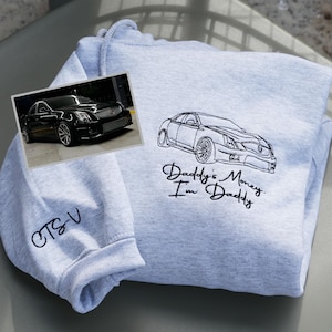 May include: Light gray hoodie with a black car outline and the text "Daddy's Money, I'm Daddy." A small photo of a black car is on the front. The sleeve has "CTS-V" embroidered in black.