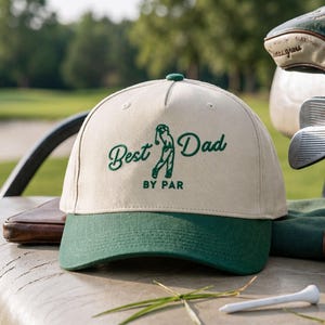 May include: A beige and green baseball cap with the text "Best Dad BY PAR" embroidered in green. The cap features a green brim and a green button on top. The image is taken outdoors, with golf clubs and a golf course in the background.
