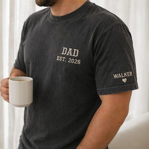 May include: A charcoal gray short-sleeve t-shirt featuring white embroidered text on the chest that reads DAD EST. 2026. The left sleeve includes the embroidered name WALKER followed by a small white heart symbol, all in a clean font.