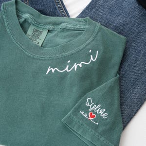 Könnte beinhalten: Ein teal Comfort Colors T-Shirt mit dem Wort "mimi" in weißer Schrift über der Brust gestickt. Der Ärmel zeigt den Namen "Sylvie" mit einem roten Herzen und dekorativen Schnörkeln.