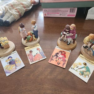 May include: Four porcelain figurines of children engaged in various activities, each paired with a miniature Good Housekeeping magazine cover. The figurines depict children with flowers, fruit, reading, and gardening, all mounted on round wooden bases.