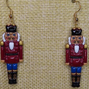 Può includere: Un paio di orecchini a gancio color oro con figure di soldatini schiaccianoci in miniatura e dettagliate. Gli schiaccianoci sono dipinti in rosso, bianco, blu e nero, con accenti dorati. Gli orecchini sono su uno sfondo beige strutturato.