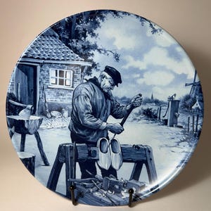 May include: A blue and white decorative plate featuring a detailed illustration of a man crafting wooden shoes. The scene includes a workshop, tools, and a rural landscape with a windmill and house.