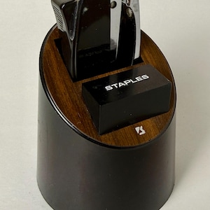 May include: A vintage stapler set featuring a black stapler and staple remover, along with a box of staples, housed in a dark wood and black holder. The word "STAPLES" is visible on the staple box.