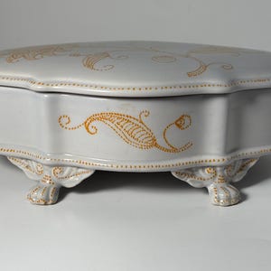 Op de afbeelding: Een hartvormige, lichtgrijze keramische sieradendoos met een deksel. De doos heeft sierlijke, goudkleurige bloemmotieven en staat op vier decoratieve voetjes. De randen zijn geaccentueerd met kleine gouden stippen.