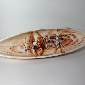 Puede incluir: Plato de cerámica ovalado con base crema y diseño en espiral marrón. El plato presenta una ilustración negra de una cabaña, árboles y montañas. Una pieza decorativa para servir aperitivos o exhibir como arte.