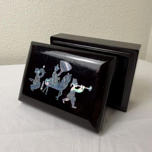 Può includere: Una scatola rettangolare laccata nera con coperchio, con un intarsio in madreperla di figure a cavallo e che suonano strumenti. La scatola è aperta, rivelando l'interno. La finitura lucida riflette la luce.