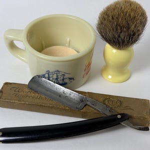 Op de afbeelding: Een vintage scheerset met een crèmekleurige mok met een scheepsillustratie, een scheerkwast en een recht scheermes met een zwarte handgreep. Het scheermes ligt op een kartonnen doos met tekst. De set is ontworpen voor de verzorging.