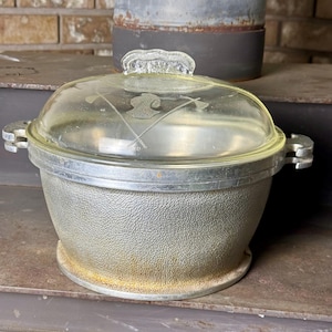 Puede incluir: Una olla redonda de aluminio con exterior texturizado y dos asas laterales. La olla incluye una tapa de cristal transparente con un diseño grabado de una figura central flanqueada por dos hachas cruzadas. La base de metal muestra ligera oxidación.