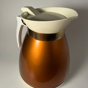 May include: A vintage copper-coloured insulated jug with a white handle, lid, and base. The jug has a gold-coloured band around the middle. The lid has a spout for pouring. The jug is designed to keep liquids hot or cold.