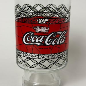 Può includere: Un bicchiere in vetro trasparente con una fascia rossa che riporta la scritta bianca Enjoy Coca-Cola e un simbolo di marchio registrato nero. Il design presenta un motivo a reticolo geometrico nero sopra e sotto la sezione rossa.