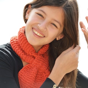 May include: A young person with long brown hair is wearing a black top with a bright orange knitted scarf. The scarf is tied in a loose knot around the neck. The person is smiling and looking at the camera.