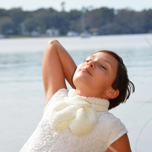 May include: A young person wearing a white crocheted scarf and a white lace top, with their eyes closed and head tilted back, relaxing outdoors by a body of water.