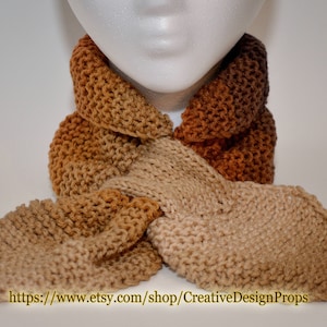 May include: A brown, beige, and tan knitted scarf with a textured pattern. The scarf is folded in half and tied around a mannequin's neck. The scarf is made of soft yarn and has a warm, cozy feel.