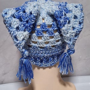 May include: A handmade crochet hat in shades of blue and white. The hat features a textured pattern and two tassels hanging from the sides. The hat is displayed on a mannequin head.