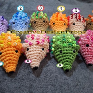 May include: A collection of ten handmade crochet hedgehogs in various colors, including purple, blue, brown, orange, pink, and green. Each hedgehog features bead accents and black button eyes. The text "CreativeDesignProps" is visible.