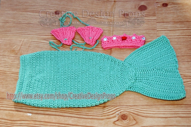 May include: A crocheted mermaid tail costume for a baby or toddler. The tail is teal green and has a matching headband with a red jewel. The set also includes a pink crocheted bikini top.