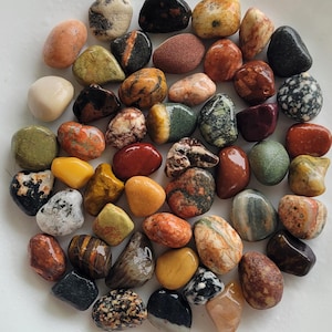 Op de afbeelding: Een verzameling van talloze kleine, gladde, gepolijste rivierstenen in diverse kleuren, waaronder tinten rood, oranje, geel, groen, zwart, wit en bruin, met uiteenlopende natuurlijke texturen, spikkels en minerale patronen.