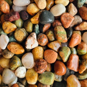May include: A collection of smooth, polished river stones in a variety of colors including shades of orange, yellow, green, brown, white, and grey. The stones are rounded and vary in size, creating a dense, textured surface of natural pebbles.