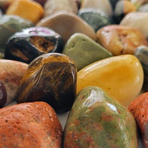 Puede incluir: Una colección de piedras pulidas y lisas de diversas formas y tamaños. El surtido incluye rocas en tonos rojo intenso, amarillo dorado, marrón oscuro, verde bosque, negro y crema, todas con un acabado brillante y reflectante.