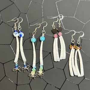 May include: A collection of handmade earrings with various designs. Some earrings feature blue and white beads, turquoise beads, or heart-shaped beads. Others have colorful gemstone accents and silver-tone bow charms. The earrings have long, white, shell-like pendants.