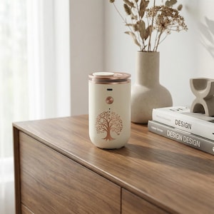 May include: A cream-coloured essential oil diffuser with a rose gold top and accents. The diffuser has a tree design on the front. It sits on a wooden surface next to a vase with dried flowers and books.