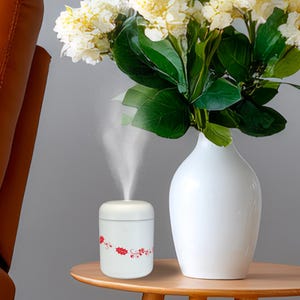 Könnte beinhalten: Ein weißer Diffusor für ätherische Öle mit rotem Blumendesign, der einen feinen Nebel abgibt. Er steht auf einem Holztisch neben einer weißen Vase mit weißen Blumen und grünen Blättern. Der Hintergrund ist ein neutrales Grau.