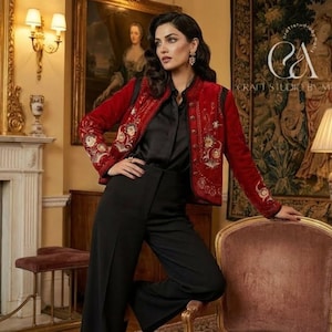 May include: A woman in a red embroidered jacket, black blouse, and black trousers. The jacket has gold and white floral embroidery. The setting includes a fireplace, paintings, and a tapestry. The logo "C&A Craft Studio by Malik" is displayed.