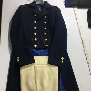Peut inclure: Un manteau en laine bleu marine avec des boutons dor&eacute;s, associ&eacute; &agrave; un pantalon cr&egrave;me. Le manteau pr&eacute;sente un design crois&eacute; et une silhouette longue et ajust&eacute;e. L'ensemble est pr&eacute;sent&eacute; sur une surface blanche.