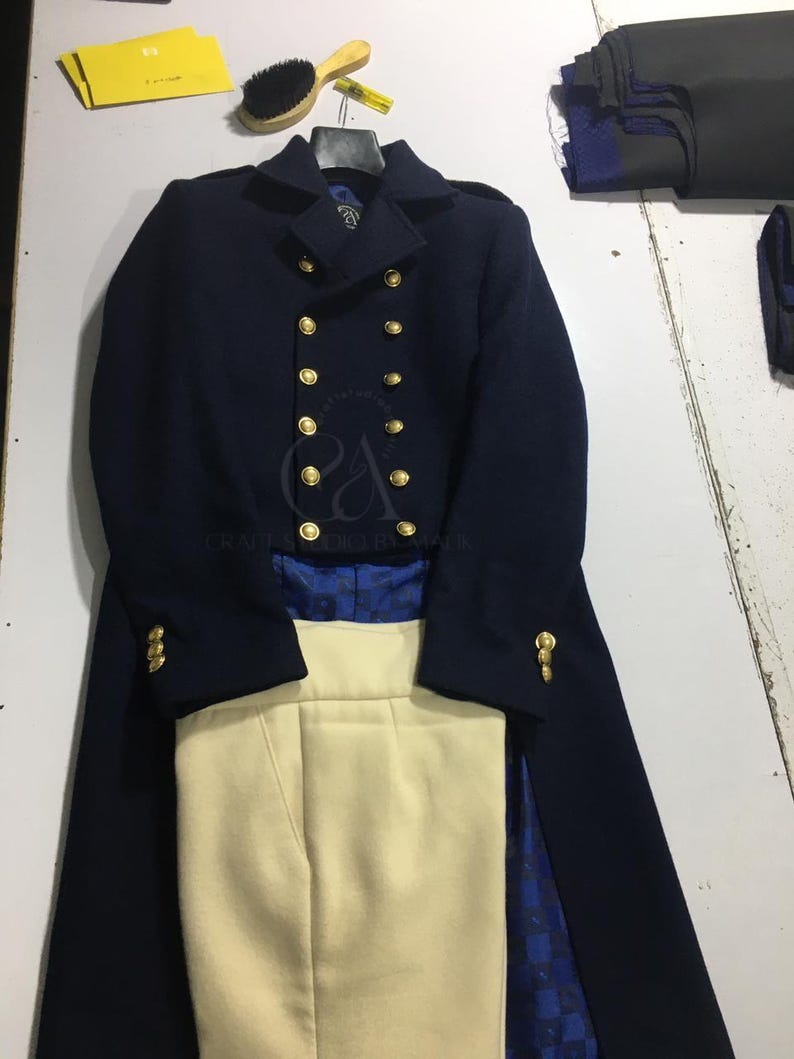 Peut inclure: Un manteau en laine bleu marine avec des boutons dor&eacute;s, associ&eacute; &agrave; un pantalon cr&egrave;me. Le manteau pr&eacute;sente un design crois&eacute; et une silhouette longue et ajust&eacute;e. L'ensemble est pr&eacute;sent&eacute; sur une surface blanche.