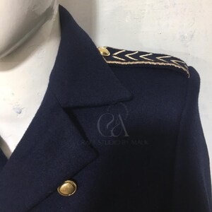 Peut inclure: Gros plan sur un manteau bleu marine avec des boutons dor&eacute;s et des d&eacute;tails aux &eacute;paules. Le manteau pr&eacute;sente une conception crois&eacute;e et un motif de chevrons dor&eacute;s sur les &eacute;paules. Le texte "Craft Studio by Malik" est visible.