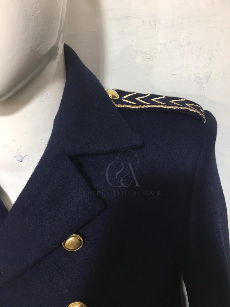Peut inclure: Gros plan sur un manteau bleu marine avec des boutons dor&eacute;s et des d&eacute;tails aux &eacute;paules. Le manteau pr&eacute;sente une conception crois&eacute;e et un motif de chevrons dor&eacute;s sur les &eacute;paules. Le texte "Craft Studio by Malik" est visible.
