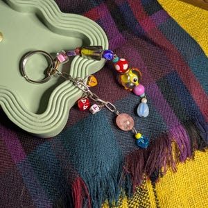 May include: A colorful beaded keychain with a silver ring. The keychain features a variety of beads, including clear, pink, blue, and red, along with a small charm. The background includes a green tray and a plaid scarf.