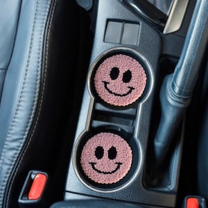 May include: Two pink car coasters with a smiley face design. The coasters are round and fit inside a car's cup holders. The smiley faces have black eyes and a black curved mouth. The coasters are made of a textured material.