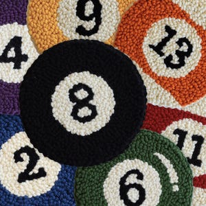 May include: A collection of circular punch needle coasters featuring the designs of billiard balls. Each coaster displays a unique number in black, including 2, 4, 6, 8, 9, 11, and 13, set against a white circular center and vibrant colored borders.