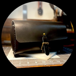 May include: A black leather bag with a flap closure and a brass buckle. The bag has a rectangular shape and a stitched edge. The bag is sitting on a white surface.