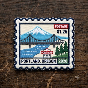 May include: A rectangular embroidered patch shaped like a postage stamp featuring a blue mountain, a dark gray suspension bridge, a river, and green pine trees. Text includes POSTAGE $1.25, PORTLAND, and 2026. The border is white with navy blue scalloped edges.