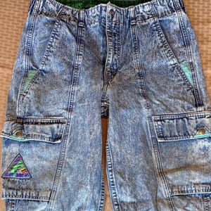 May include: Acid wash denim cargo pants with multiple pockets and a high waist. The pants feature a colorful triangle patch on one pocket and teal and green accents. The waistband is elastic and the pants have a relaxed fit.