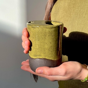 Può includere: Una tazza in ceramica fatta a mano con un design unico. La metà superiore è verde oliva macchiato, mentre la parte inferiore è marrone scuro. La tazza ha una parte superiore curva con una piccola apertura. La tazza è tenuta nelle mani di una persona.