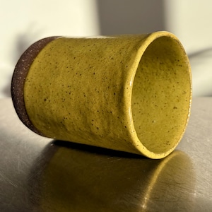 May include: A handcrafted ceramic mug in a warm yellow colour with a speckled texture. The rim is a contrasting dark brown. The mug is lying on its side, showing its interior and the reflective surface beneath.