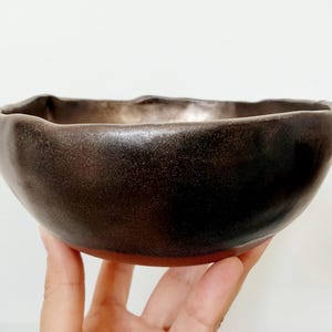 May include: A handmade ceramic bowl with a dark brown exterior and a lighter interior. The bowl has a rough, uneven rim and a reddish-brown base. The bowl is held in a hand.