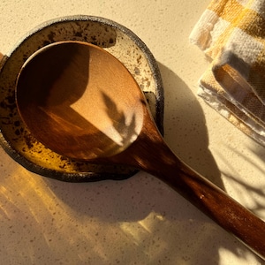 May include: A wooden spoon rests on a ceramic spoon rest with a speckled design. The spoon and rest are brown and tan. A folded yellow and white striped kitchen towel is in the background.