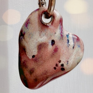 May include: A heart-shaped pendant with a speckled, glazed finish. The pendant features a mix of colours, including shades of pink, white, and dark spots. A silver-coloured metal ring is attached at the top for hanging.
