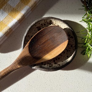 May include: A wooden spoon rests on a speckled ceramic spoon rest. A yellow and white checkered kitchen towel is in the upper left corner. Fresh rosemary sprigs are in the upper right corner. The spoon is dark brown.