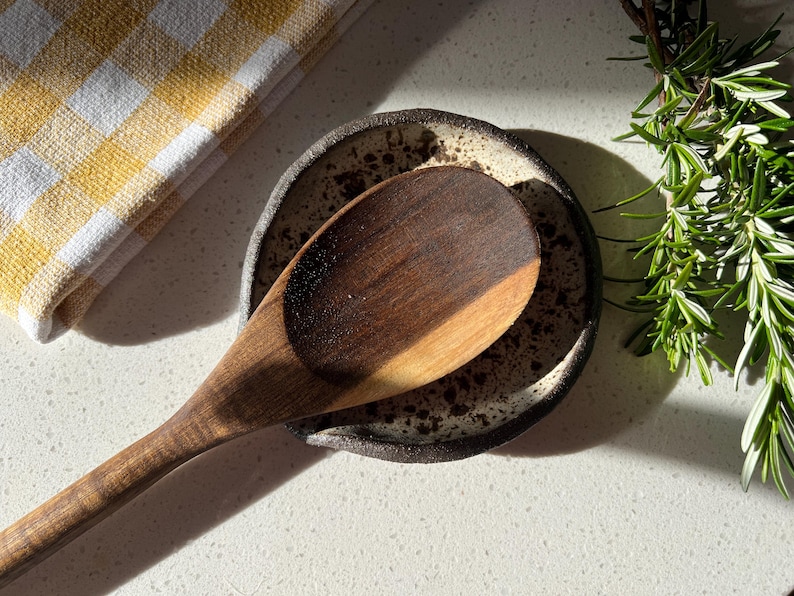 May include: A wooden spoon rests on a speckled ceramic spoon rest. A yellow and white checkered kitchen towel is in the upper left corner. Fresh rosemary sprigs are in the upper right corner. The spoon is dark brown.