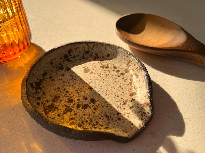 May include: A speckled ceramic spoon rest with a dark rim and a wooden spoon. The spoon rest is off-white with brown speckles. An orange glass is visible in the background. The items are on a light-colored surface.