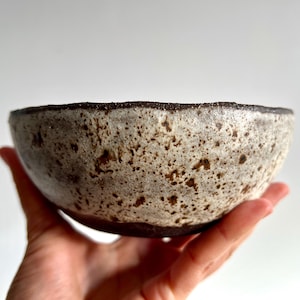 Puede incluir: Un cuenco de cerámica hecho a mano con un exterior moteado blanquecino y un borde marrón oscuro. El cuenco se sostiene en una mano, mostrando su superficie texturizada y su forma orgánica. El interior del cuenco no es visible.