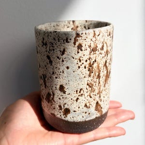 May include: A cylindrical ceramic cup featuring a speckled off-white and cream glaze on the upper portion, transitioning to a solid dark brown base. The surface has a textured appearance with irregular brown spots scattered across the body.