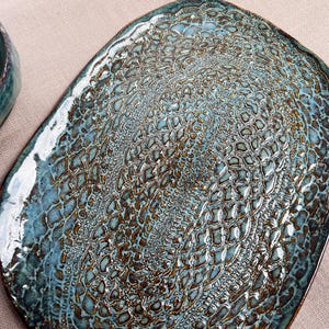 May include: A handmade ceramic serving platter with a unique textured surface. The oval-shaped platter features a teal and brown glaze, creating a striking contrast. The intricate pattern resembles a lace design, adding a touch of elegance to the piece. A matching bowl is visible in the background.