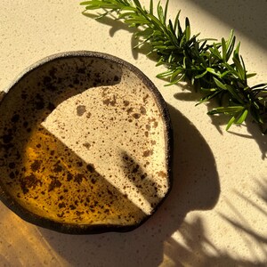 May include: A small, handmade ceramic dish with a speckled, off-white interior and a dark brown rim. The dish is partially filled with a golden-brown glaze. A sprig of fresh rosemary rests nearby, casting a shadow.